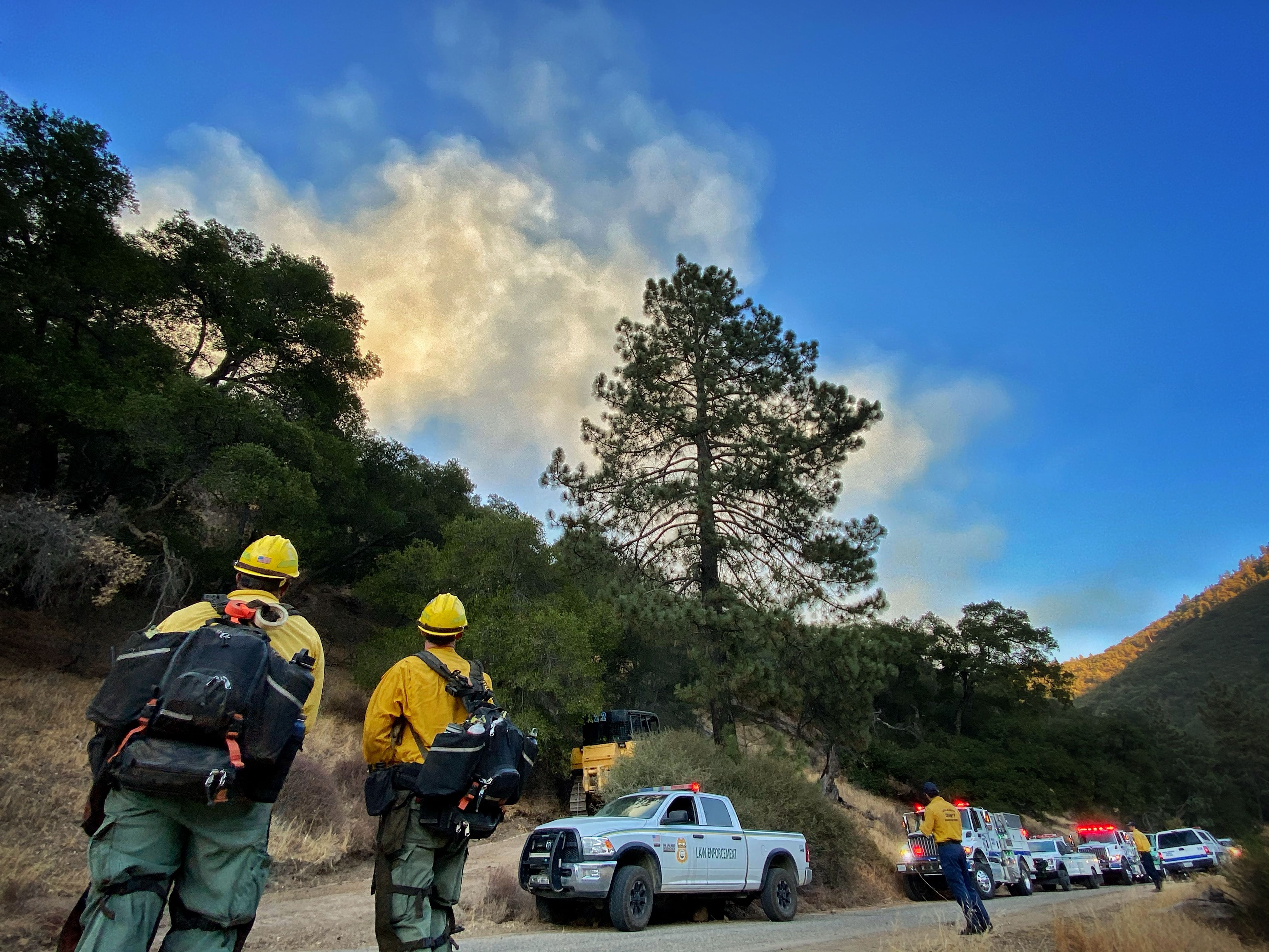 Two Santa Barbara firefighters at a staging area, with firetrucks parked against a hill and smoke in the blue sky beyond the hill.